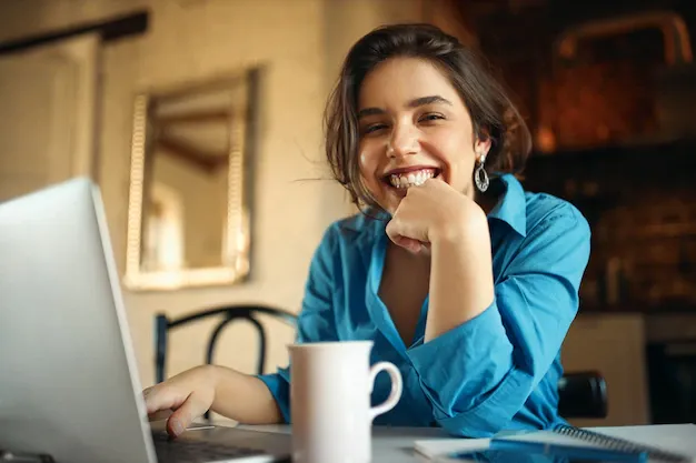 cheerful-attractive-young-woman-enjoying-distant-work-sitting-desk-using-portable-computer-drinking-coffee-pretty-female-blogger-working-from-home-uploading-video-her-channel-smiling_343059-2096.webp