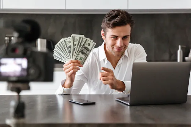 smiling-young-man-filming-his-video-blog-episode-about-new-tech-devices-while-sitting-kitchen-table-with-laptop-showing-bunch-money-banknotes_171337-5530.webp