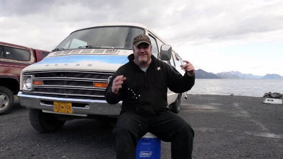Smiling man sits on cooler in front of van by the Alaska sea shore. Is he homeless or not? You can’t tell.