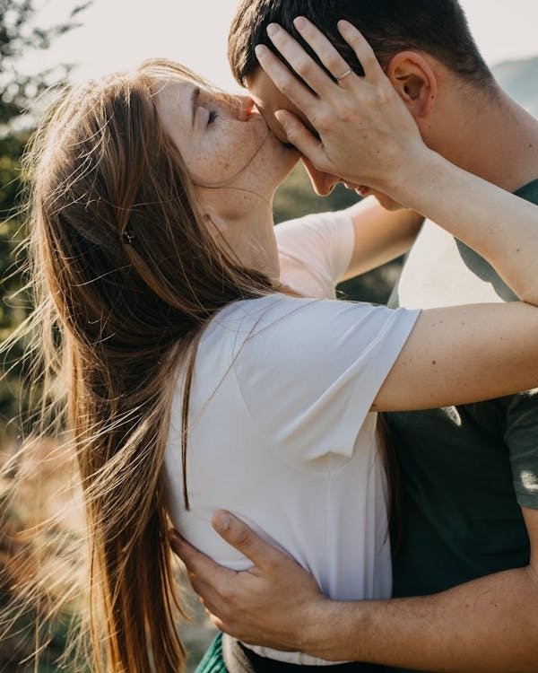 Young romantic couple standing on grassy meadow in countryside embracing while woman kissing man on forehead with tenderness: