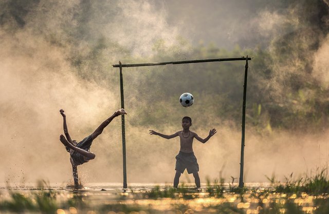2 kids playing soccer in a wet field with bamboo poles for the goal