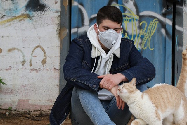 young man sitting and wearing a mask with a cat rubbing its head against his hands