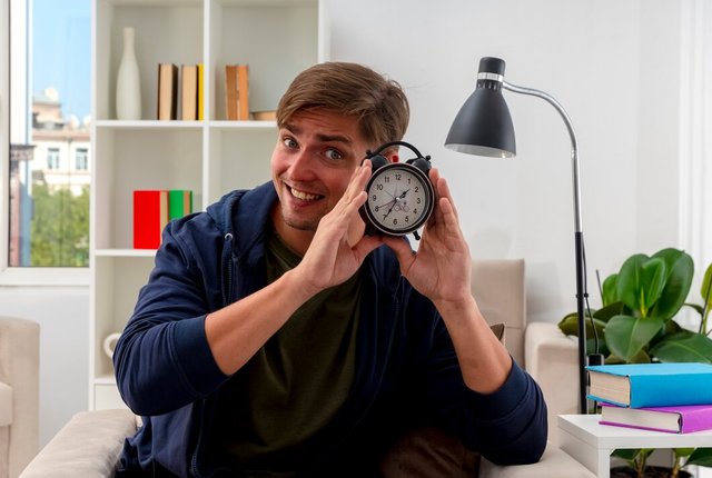 smiling-young-blonde-handsome-man-sits-armchair-holding-alarm-clock-inside-living-room_141793-70143.jpg