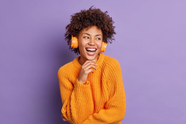 positive-dark-skinned-young-woman-listens-music-headphones-holds-chin-smiles-pleased-concentrated-aside-feels-overjoyed-wears-warm-knitted-orange-jumper_273609-45253.jpg