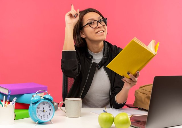 smiling-young-student-girl-wearing-glasses-sitting-desk-with-university-tools-holding-book-doing-homework-with-raised-finger-isolated-pink-wall_141793-97690.jpg