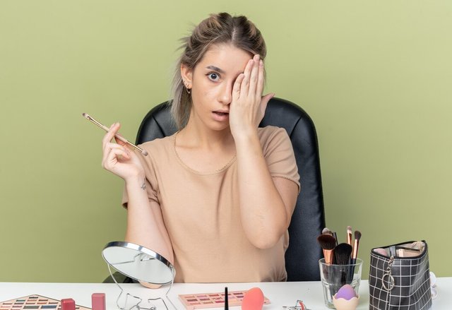 surprised-young-beautiful-girl-sitting-desk-with-makeup-tools-holding-makeup-brush-covered-eye-with-hand-isolated-olive-green-background_141793-119310.jpg