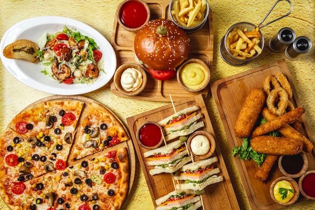 top-view-fast-food-mix-mozzarella-sticks-club-sandwich-hamburger-mushroom-pizza-caesar-shrimp-salad-french-fries-ketchup-mayo-cheese-sauces-table_141793-3998.jpg