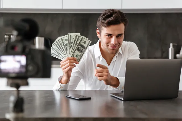 smiling-young-man-filming-his-video-blog-episode-about-new-tech-devices-while-sitting-kitchen-table-with-laptop-showing-bunch-money-banknotes_171337-5530.webp