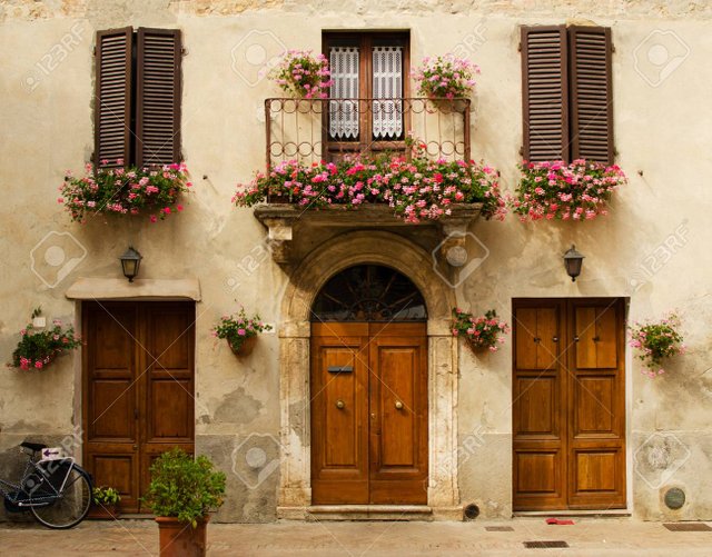 92517047-fachada-de-la-casa-antigua-en-pienza-con-ventanas-balcón-con-flores-puertas-de-madera-plantas-toscana-i.jpg