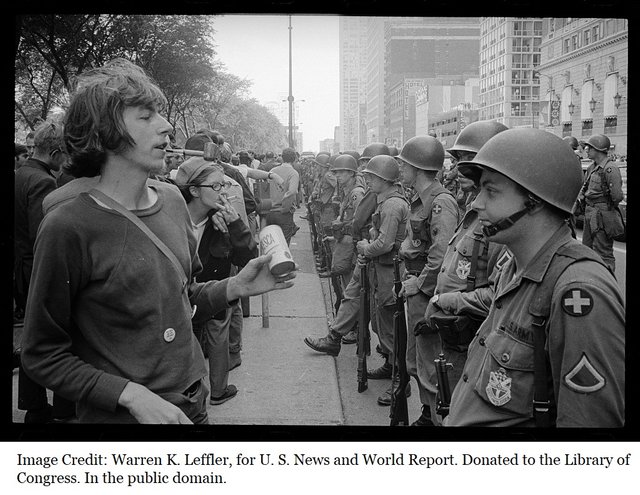 Young_'hippie'_standing_in_front_of_a_row_of_National_Guard_soldiers,_across_the_street_from_the_Hilton_Hotel_at_Grant_Park,_at_the_Democratic_National_Convention_in_Chicago,_August_26,_1968.jpg