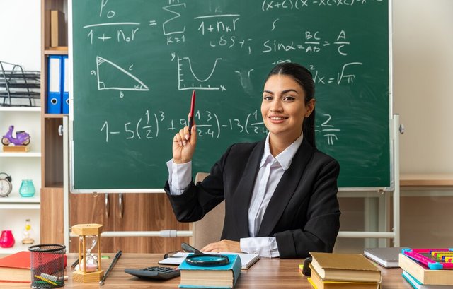 impressed-young-female-teacher-sits-table-with-school-tools-raising-pen-classroom_141793-106366.jpg