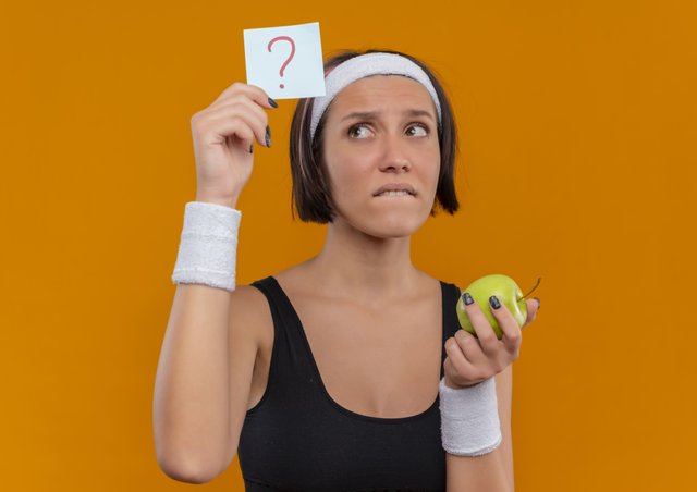 young-fitness-woman-sportswear-with-headband-showing-reminder-paper-with-question-mark-holding-green-apple-looking-paper-confused-standing-orange-wall (1).jpg