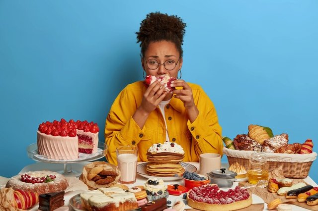 gluttony-overeating-concept-upset-crying-ethnic-woman-eats-piece-cake-reluctantly-sits-table-with-many-desserts-isolated-blue-wall_273609-39244.jpg