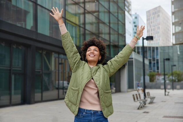 horizontal-shot-happy-carefree-woman-with-curly-hair-keeps-arms-raised-up-enjoys-life-poses-during-daytime-city-wears-jacket-jeans-looks-overhead-has-cheerful-expression_273609-55849.jpg