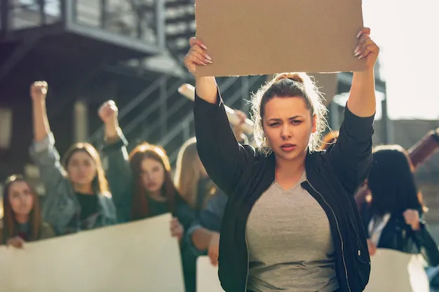 young-woman-with-blank-poster-front-people-protesting-about-women-s-rights-equality-street-meeting-about-problem-workplace-male-pressure-domestic-abuse-harassment-copyspace_489646-7753.webp