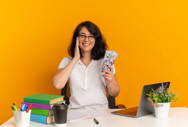 young-smiling-pretty-caucasian-schoolgirl-wearing-glasses-sits-desk-with-school-tools-puts-hand-face-holds-money-isolated-orange-space-with-copy-space_141793-60006.jpg