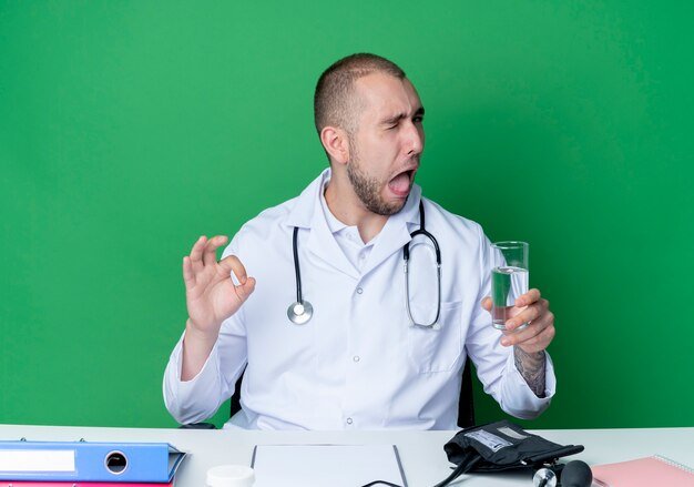 confident-young-male-doctor-wearing-medical-robe-stethoscope-sitting-desk-with-work-tools-holding-glass-water-winking-doing-ok-sign-isolated-green_141793-77185.jpg