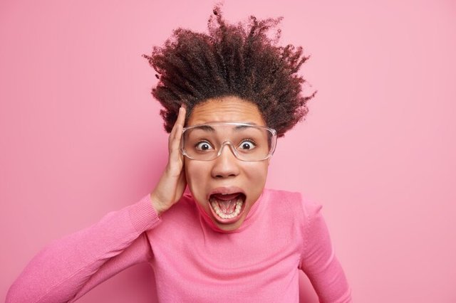 very-scared-emotional-crazy-curly-woman-shouts-loudly-keeps-hand-head-mouth-wide-opened-stares-bugged-eyes-through-transparent-glasses-terrified-by-awful-scene_273609-50674.jpg