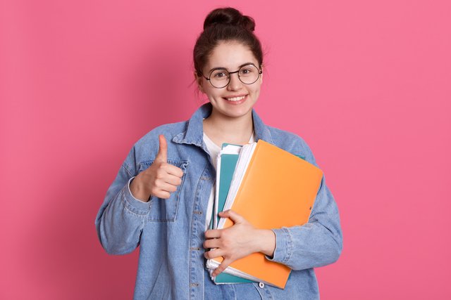 young-student-woman-wearing-denim-jacket-eyeglasses-holding-colorful-folders-showing-thumb-up-pink.jpg