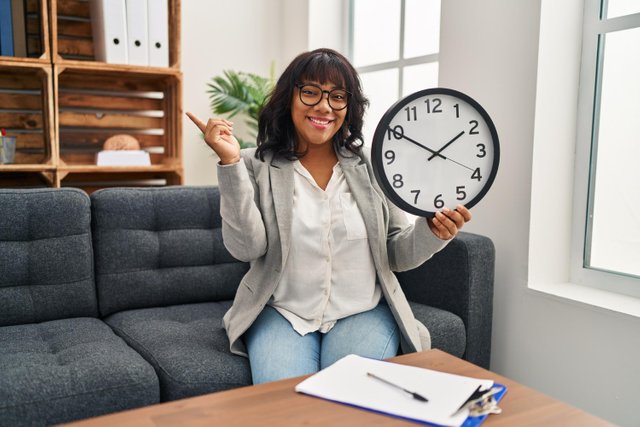 hispanic-woman-working-therapy-office-holding-clock-smiling-happy-pointing-with-hand-finger-side.jpg