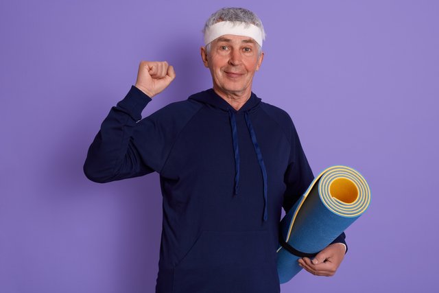 elderly-man-clenching-fist-holding-yoga-mat-wearing-sport-clothing-headband-posing-isolated-lilac.jpg