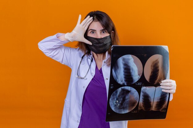 middle-aged-doctor-wearing-white-coat-black-protective-facial-mask-with-stethoscope-holding-x-ray-lungs-looking-surprised-with-hand-near-head-orange-wall_141793-13275.jpg