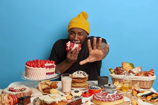 hungry-displeased-plump-afro-man-keeps-palm-forward-camera-bites-huge-piece-creamy-cake-gets-much-calories-surrounded-with-tasty-baked-products_273609-34541.jpg