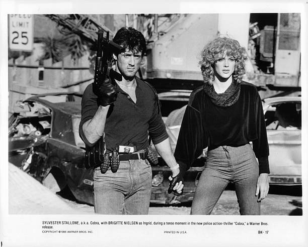 sylvester-stallone-with-a-gun-in-one-hand-is-holding-brigitte-hand-picture-id128010308.jpeg