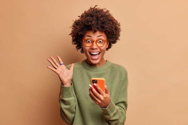 overjoyed-excited-woman-with-afro-hair-raises-palm-has-eyes-full-happiness-after-receiving-excellent-news-holds-mobile-phone-wears-spectacles-optical-glasses-isolated-brown-wall_273609-44115.jpg