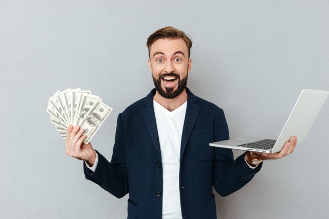 happy-surprised-bearded-man-busines-clothes-holding-money-laptop-computer-while-looking-camera-gray.jpg