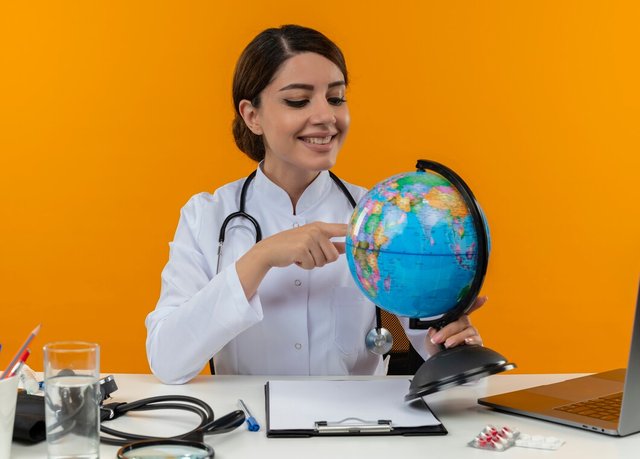 smiling-young-female-doctor-wearing-medical-robe-with-stethoscope-sitting-desk-work-computer-with-medical-tools-holding-putting-finger-globe-isolation-yellow-background_141793-37961.jpg