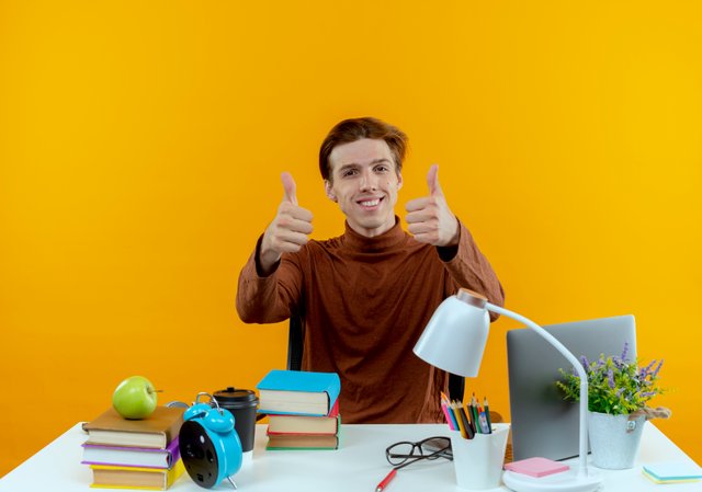 smiling-young-student-boy-sitting-desk-with-school-tools-his-thumbs-up-isolated-yellow-wall.jpg