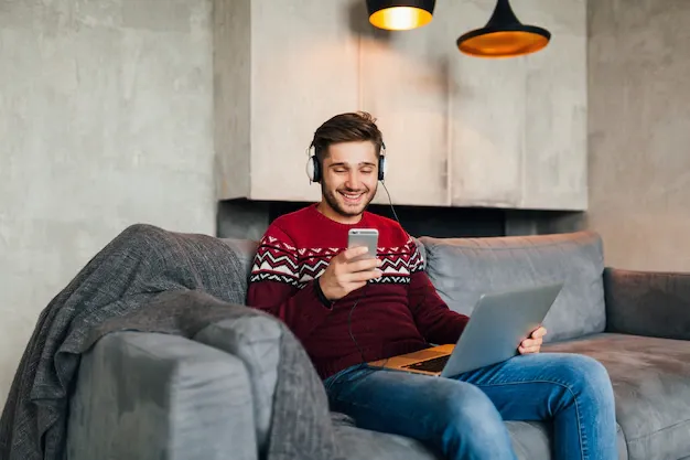 young-attractive-man-sofa-home-winter-with-smartphone-headphones-listening-music-wearing-red-knitted-sweater-working-laptop-freelancer_285396-2726.webp