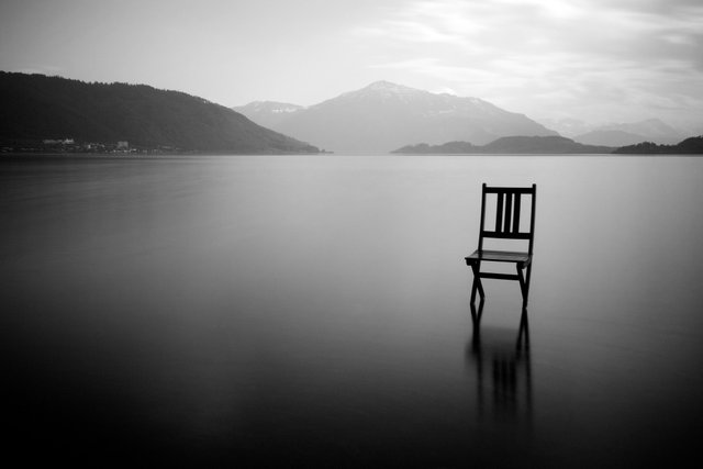 a wooden chair rests on the smooth surface of still water; mountains rest in the misty background