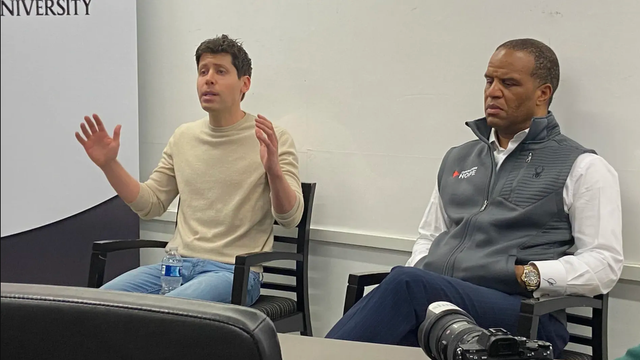 A man speaks with his arms outstretched in a classroom while a man seated next to him listens intently