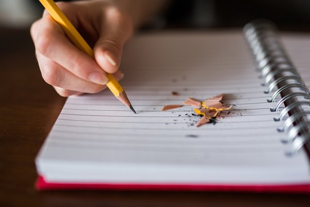 A person writing with a pencil in a notebook with pencil shavings on it
