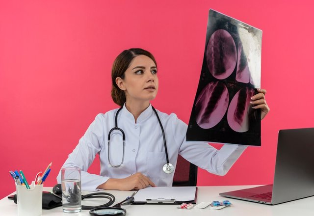 pleased-young-female-doctor-wearing-medical-robe-with-stethoscope-sitting-desk-work-computer-with-medical-tools-holding-looking-x-ray-with-copy-space_141793-37718.jpg