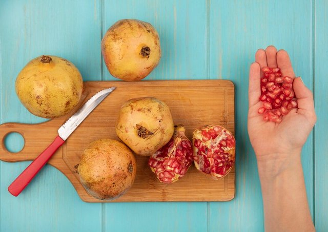 top-view-female-hand-holding-pomegranate-seeds-with-whole-pomegranates-wooden-kitchen-board-with-knife-blue-surface_141793-80513.jpg