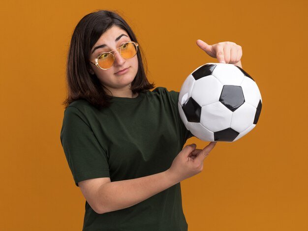 Free photo impressed young pretty caucasian girl in sun glasses holding and looking at ball isolated on orange wall with copy space