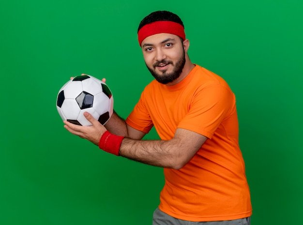 Free photo pleased young sporty man wearing headband and wristband holding out ball at side isolated on green background
