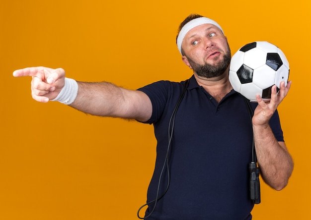 Free photo surprised adult slavic sporty man with jumping rope around neck wearing headband and wristbands holding ball and pointing at side isolated on orange wall with copy space