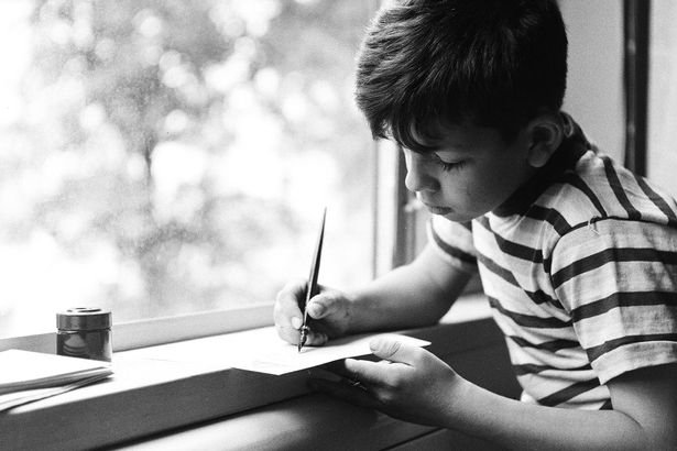 A-young-boy-in-a-striped-shirt-sits-by-a-window-and-writes-with-a-quill-pen.jpg