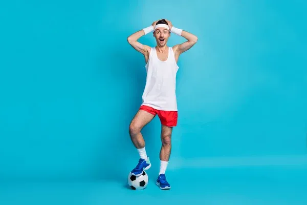 Full body portrait of impressed young man arms on head open mouth leg on ball isolated on blue color background — Stock Photo, Image