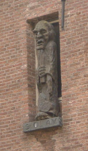 Bild der Olav–Statur in der Niesche einer Backsteinmauer. Olav hat den Mund geöffnet uns schaut sehr grimmig. er hält eine Art Schriftrolle senkrecht vor den Körper.