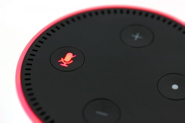 An Amazon Alexa digital assistant: a circular black device with a red lit ring around it, featuring power, microphone/mute, and volume increase/decrease buttons.