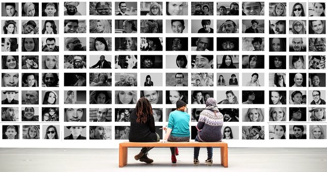 Three women sitting on a bench and looking at a wall covered in black and white images of various people. A mind-reading AI might be able to describe what each woman was seeing.