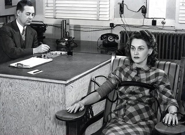 A black-and-white photo of a polygraph test being administered by an older man to a young woman in a checkered dress in an office, circa 1945.