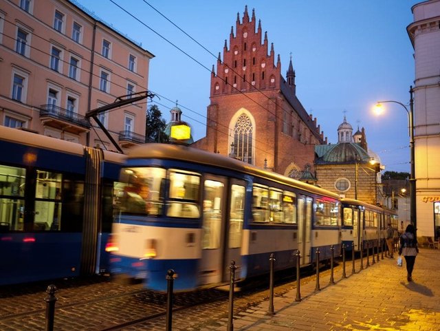 A blue and white tram, blurry, going down the street in Krakow, a spiky church behind it.