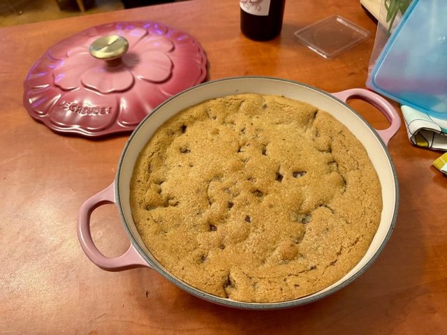 A giant cookie in a cast-iron skillet, with its bright pink flower-shaped lid to the side.